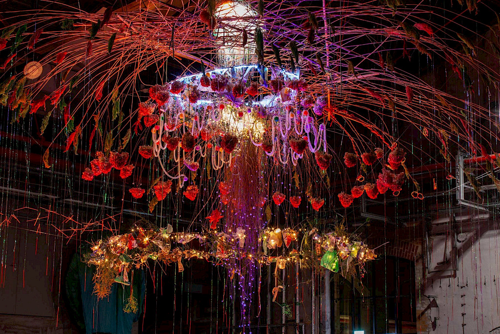 Foto einer Installation von Renate Liebel und Justyna Koeke.  Die Installation sieht aus wie ein riesiger Kronleuchter. Viele getrocknete Blumen und andere Pflanzen, Schaschlik-St&auml;bchen und M&uuml;ll sind in Neonfarben bemalt. Sie h&auml;ngen mit Lichterketten von der Decke.