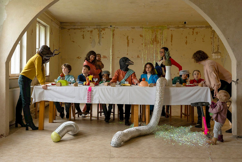 Installation von Maternal Fantasies. An einem langen Tisch mit wei&szlig;er Tischdecke sitzen einige Kinder. Drei weiblich gelesene Personen und eine Person mit Tierkopf stehen bei ihnen. In der Mitte sitzt eine Person mit einem Vogelkopf. Auf und vor dem Tisch sind Brot, Obst und Spielsachen verteilt. Die Fotografie erinnert an das letzte Abendmahl.