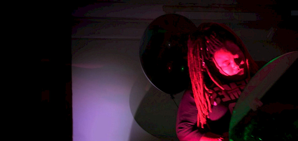 Die Fotografie zeigt die kongolesische Performerin Orakle Ngoy bei einer Live-Performance im Theater Rampe. Sie h&auml;lt einen gro&szlig;en Spiegel in der Hand, ihr Gesicht ist in rotes Licht getaucht. Im Hintergrund schwebt ein gro&szlig;er schwarzer Ballon &ndash; dieser ist Teil einer Installation der K&uuml;nstlerin Marie Lienhard. Die Szene findet in einem abgedunkelten Raum mit farbigen Lichtprojektionen statt und verbindet Performancekunst mit Installation.